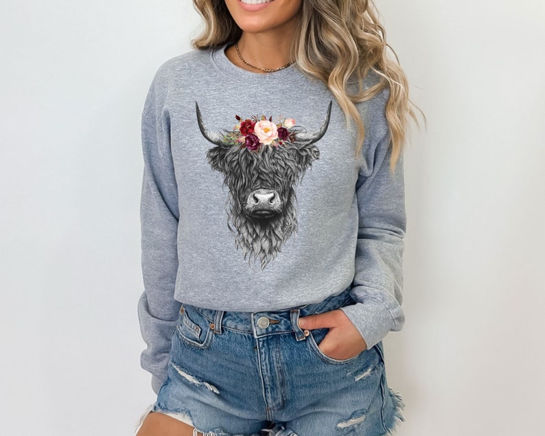 Puede incluir: Una sudadera gris con una ilustraci&oacute;n en blanco y negro de una vaca de las tierras altas con una corona de flores. Las flores son rojas, rosas y blancas.