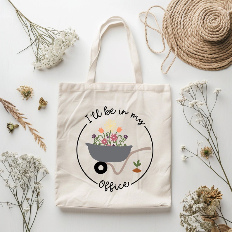 May include: A white canvas tote bag with a black circle and the text "I'll be in my office" inside the circle. The circle also contains a gray wheelbarrow with colorful flowers inside.