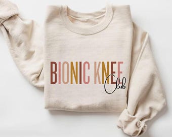May include: Cream-colored sweatshirt with the text "BIONIC KNEE Club" in brown, gold, and pink lettering. The word "Club" is in a cursive font. The sleeves are folded up, and the sweatshirt is folded.