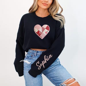 May include: Black sweatshirt with a patchwork heart design in shades of pink, red, and white. The sleeve features the name "Sophia" in a cursive font. The sweatshirt is paired with blue jeans.