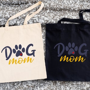 May include: Two tote bags with the text "Dog Mom" in yellow letters and a blue paw print with a red heart in the center. One bag is white and the other is black.
