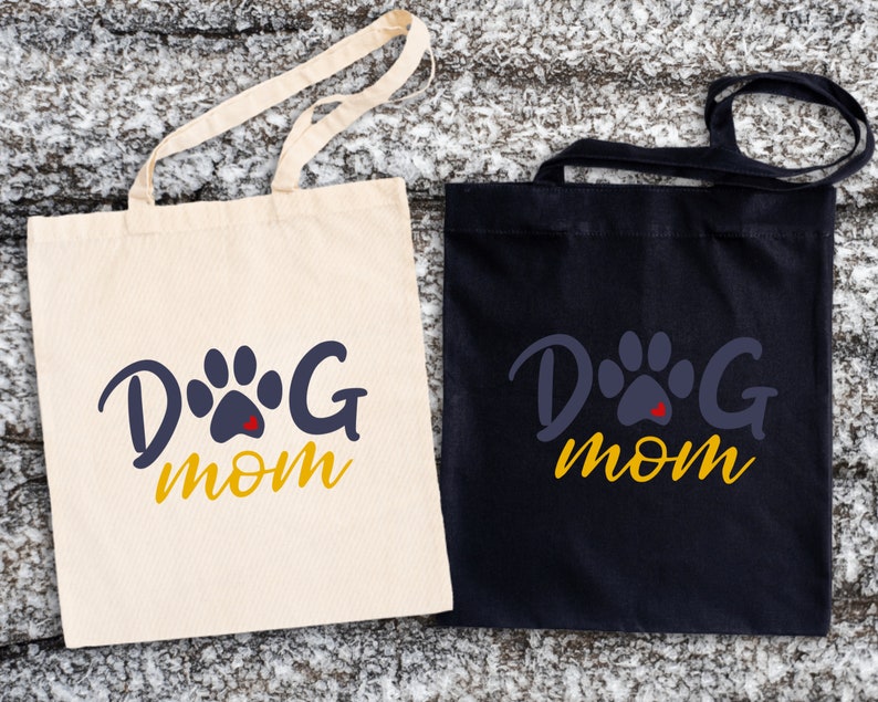 May include: Two tote bags with the text "Dog Mom" in yellow letters and a blue paw print with a red heart in the center. One bag is white and the other is black.