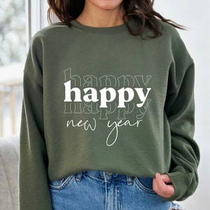 May include: An olive green sweatshirt with the words "happy new year" in white. The word "happy" is stacked three times, with the middle word in a bold font. The words "new year" are written in a cursive font.