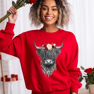 Puede incluir: Una sudadera roja con una ilustraci&oacute;n en blanco y negro de una vaca de las tierras altas con una corona de flores. La vaca tiene una melena larga y peluda y una nariz blanca.