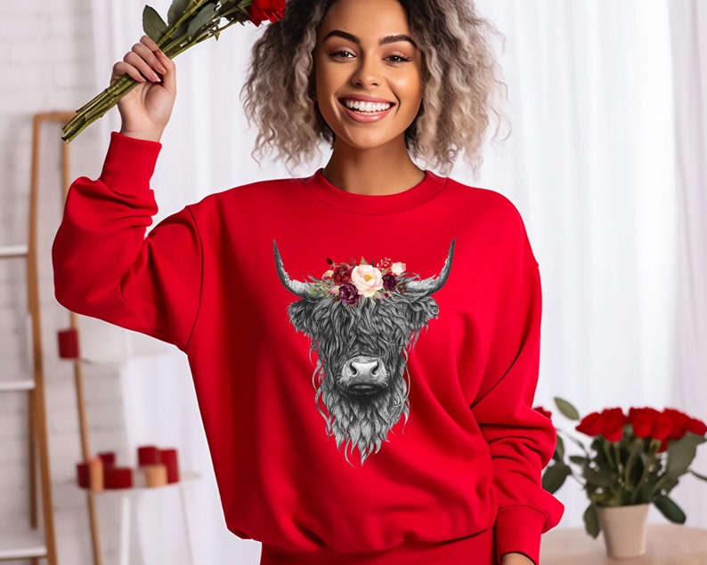 Puede incluir: Una sudadera roja con una ilustraci&oacute;n en blanco y negro de una vaca de las tierras altas con una corona de flores. La vaca tiene una melena larga y peluda y una nariz blanca.