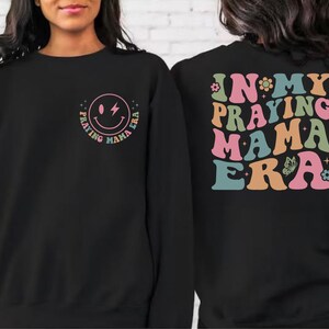 May include: Black sweatshirt with a smiley face graphic that says "Praying Mama Era" on the front and "In My Praying Mama Era" on the back. The text is in a retro font with colorful accents.