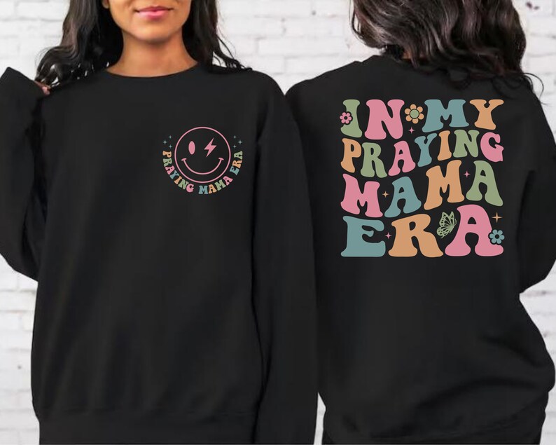 May include: Black sweatshirt with a smiley face graphic that says "Praying Mama Era" on the front and "In My Praying Mama Era" on the back. The text is in a retro font with colorful accents.