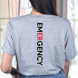 Puede incluir: Una camiseta gris jaspeada con la palabra "EMERGENCY" impresa verticalmente en negro y rojo. La camiseta tiene mangas cortas y est&aacute; hecha de un material suave.