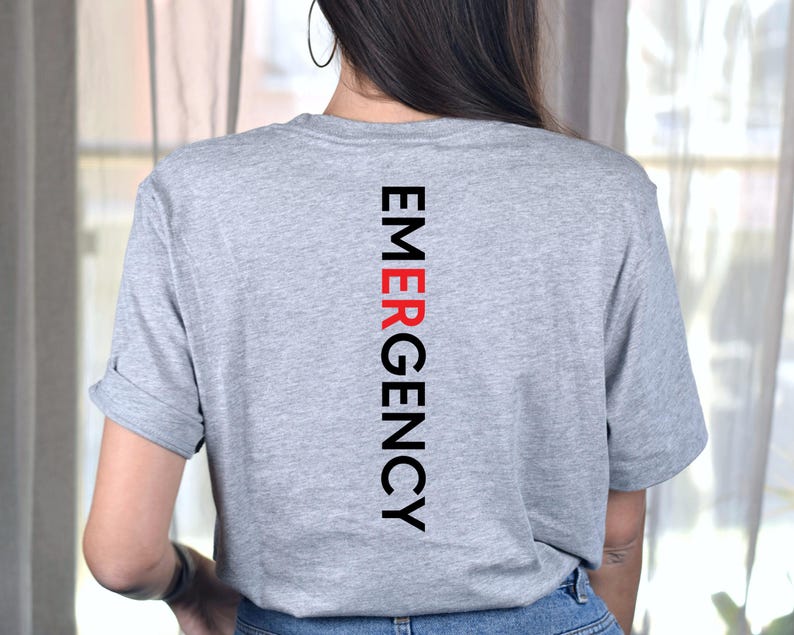 Puede incluir: Una camiseta gris jaspeada con la palabra "EMERGENCY" impresa verticalmente en negro y rojo. La camiseta tiene mangas cortas y est&aacute; hecha de un material suave.