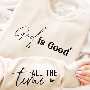 May include: A cream colored sweatshirt with the text "God is Good" and "All the Time" in black lettering. The sweatshirt has a sparkly design next to the text "God is Good".