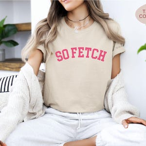 Op de afbeelding: Een beige T-shirt met de tekst "SO FETCH" in roze letters. Het shirt wordt gedragen door een persoon.