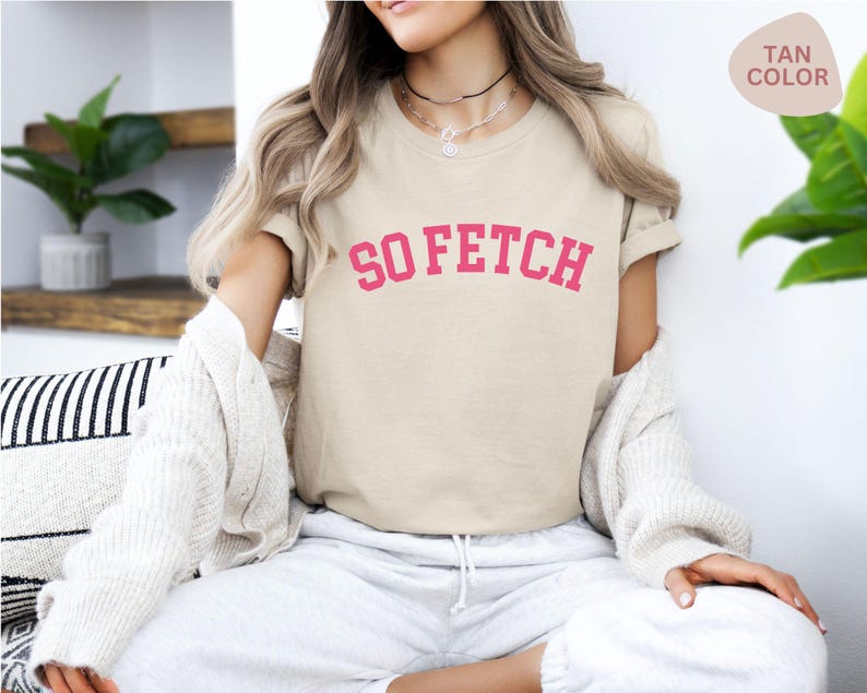 Op de afbeelding: Een beige T-shirt met de tekst "SO FETCH" in roze letters. Het shirt wordt gedragen door een persoon.