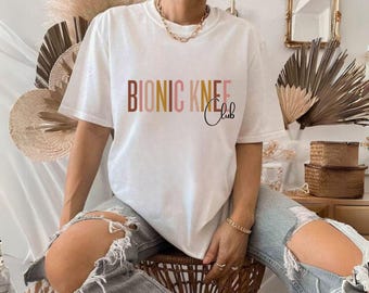 May include: A white short-sleeve t-shirt featuring the text BIONIC KNEE Club printed across the chest. The word BIONIC is in a multi-colored block font with shades of brown, orange, and yellow, while the word Club is in a black script font.