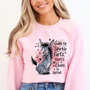 May include: A light pink sweatshirt featuring a cartoon donkey with the text "Giddy Up Sparkle Farts* there's *Chaos to Spread." The donkey is gray with a black tail and a big smile. Black stars and pink hearts surround the donkey.