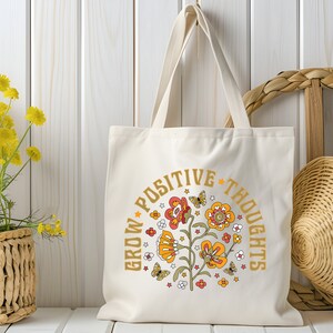 May include: A white canvas tote bag with a floral design and the text "Grow Positive Thoughts" in gold lettering.