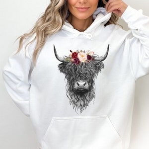 Puede incluir: Sudadera blanca con una ilustraci&oacute;n en blanco y negro de una vaca de las tierras altas con una corona de flores. Las flores son rojas, rosas y blancas.