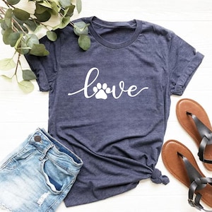 May include: A dark heather navy blue short-sleeve t-shirt featuring the word love in white cursive script, where the letter o is replaced by a white paw print graphic. The shirt is displayed alongside a pair of light blue denim shorts.