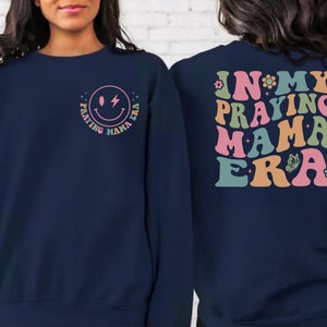 May include: Navy blue sweatshirt with a smiley face graphic and the text "Praying Mama Era" on the front. The back of the sweatshirt has the text "In My Praying Mama Era" in a retro font with colorful accents.