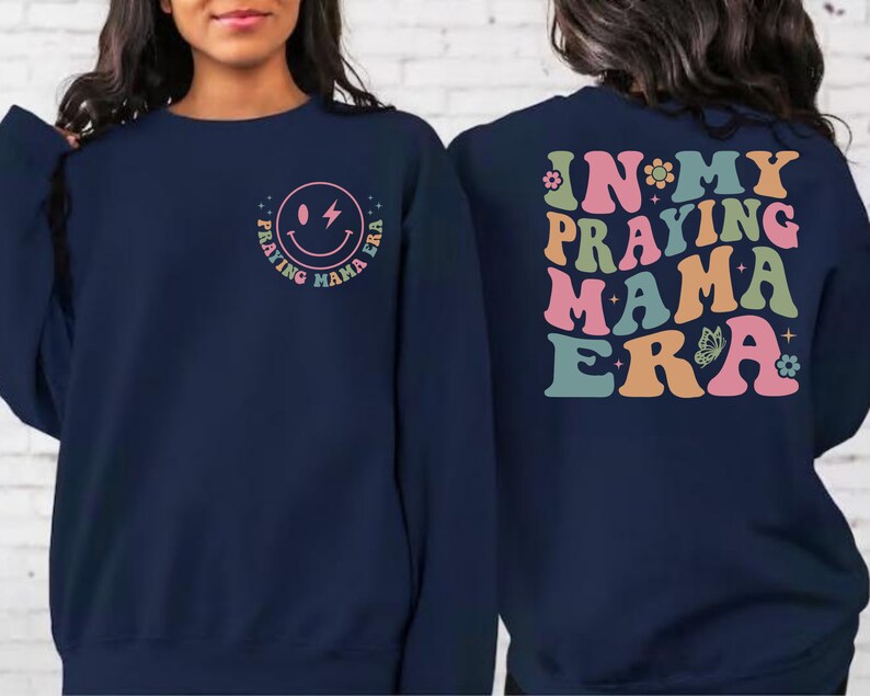 May include: Navy blue sweatshirt with a smiley face graphic and the text "Praying Mama Era" on the front. The back of the sweatshirt has the text "In My Praying Mama Era" in a retro font with colorful accents.