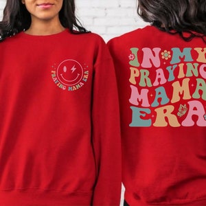 May include: Red sweatshirt with a smiley face graphic that says "Praying Mama Era" on the front and "In My Praying Mama Era" on the back.