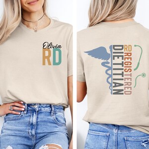 Puede incluir: Camiseta beige con el texto "Olivia RD" en negro, dorado y verde azulado. La parte trasera de la camiseta presenta un s&iacute;mbolo de caduceo con las palabras "RD Registered Dietitian" en verde azulado, marr&oacute;n y gris. La camiseta la lleva una persona.