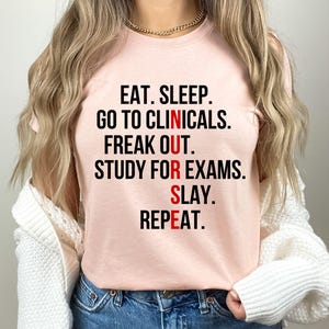 May include: A light pink t-shirt with the text "EAT. SLEEP. GO TO CLINICALS. FREAK OUT. STUDY FOR EXAMS. SLAY. REPEAT." in black and red lettering. The shirt is a crew neck style.