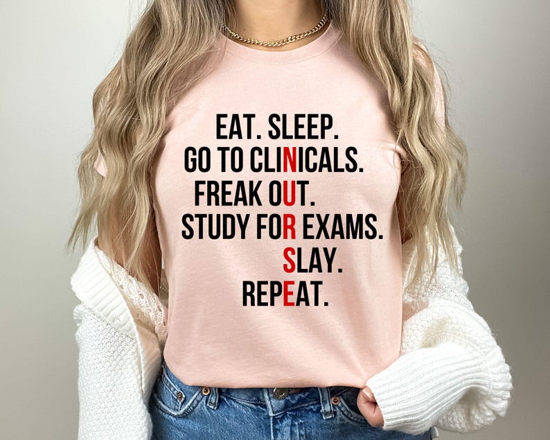 May include: A light pink t-shirt with the text "EAT. SLEEP. GO TO CLINICALS. FREAK OUT. STUDY FOR EXAMS. SLAY. REPEAT." in black and red lettering. The shirt is a crew neck style.