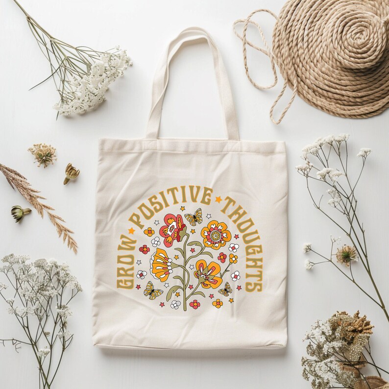 May include: A beige tote bag with a floral design and the text "Grow Positive Thoughts" in a circle around the flowers. The flowers are yellow, orange, and red with green stems and leaves. There are small white stars and butterflies scattered around the flowers.