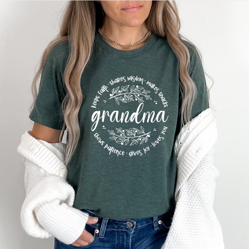 Op de afbeelding: Donkergroen T-shirt met het woord "oma" in witte letters, omcirkeld door een bloemenkrans en zinnen als "houdt geloof" en "geeft vreugde". Het T-shirt heeft een ronde hals en korte mouwen.