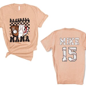 May include: Peach-colored baseball t-shirt. The front features a baseball glove, ball, and the words "BASEBALL MAMA". The back displays "MIKE 15" in a baseball-themed design.