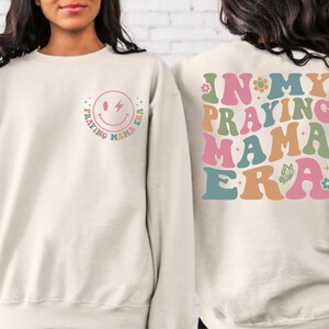 May include: A cream-colored sweatshirt with a smiley face graphic on the front that says "Praying Mama Era" in a rainbow font. The back of the sweatshirt has the same text in a larger font with a butterfly graphic.