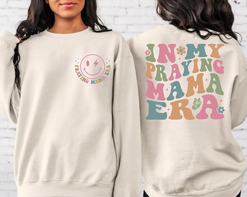May include: A cream-colored sweatshirt with a smiley face graphic on the front that says "Praying Mama Era" in a rainbow font. The back of the sweatshirt has the same text in a larger font with a butterfly graphic.