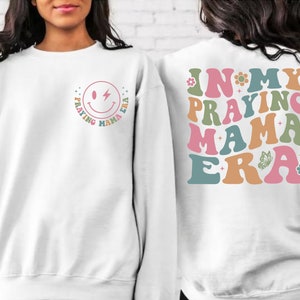 May include: White sweatshirt with a smiley face graphic on the front and the text "In My Praying Mama Era" on the back. The text is in a retro font with colorful accents.