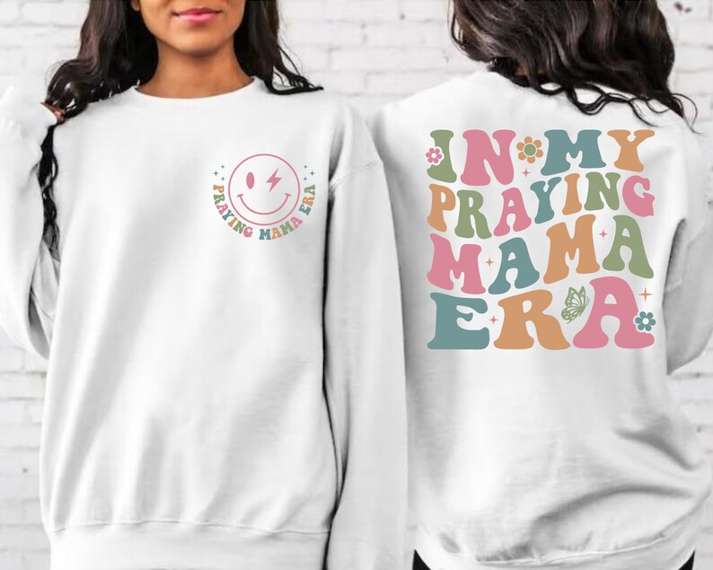 May include: White sweatshirt with a smiley face graphic on the front and the text "In My Praying Mama Era" on the back. The text is in a retro font with colorful accents.