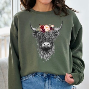 Puede incluir: Una sudadera verde con una ilustraci&oacute;n en blanco y negro de una vaca de las tierras altas con una corona de flores. Las flores son rosas, rojas y blancas.