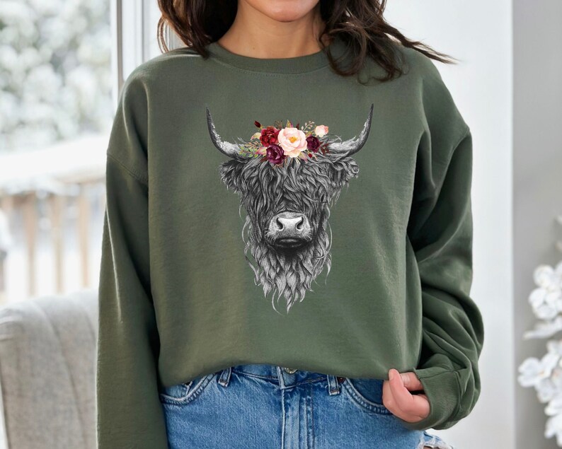 Puede incluir: Una sudadera verde con una ilustraci&oacute;n en blanco y negro de una vaca de las tierras altas con una corona de flores. Las flores son rosas, rojas y blancas.