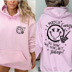 Puede incluir: Sudadera rosa con capucha con una cara sonriente negra y un gr&aacute;fico de mano de esqueleto en la parte delantera y el texto "I Match Energy So How We Gon' Act Today?" en la parte posterior.