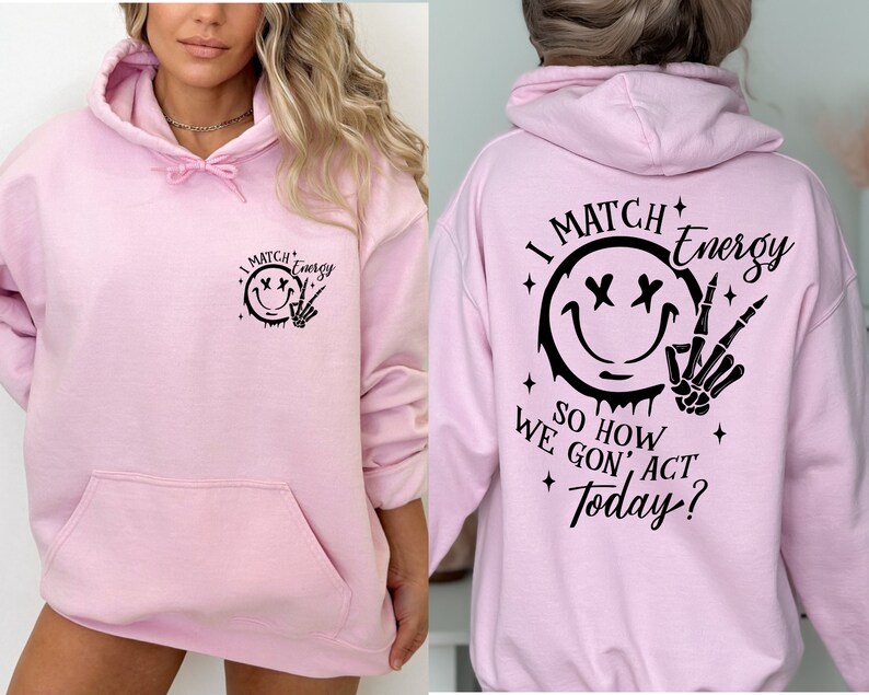 Puede incluir: Sudadera rosa con capucha con una cara sonriente negra y un gr&aacute;fico de mano de esqueleto en la parte delantera y el texto "I Match Energy So How We Gon' Act Today?" en la parte posterior.