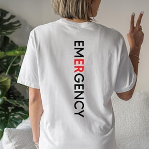 Puede incluir: Camiseta blanca con la palabra "EMERGENCY" impresa verticalmente en negro y rojo. La camiseta tiene mangas cortas y un corte relajado. El texto est&aacute; centrado en la parte trasera de la camiseta.