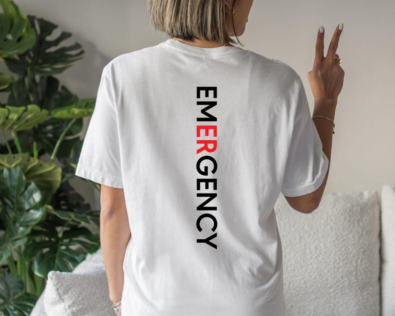 Puede incluir: Camiseta blanca con la palabra "EMERGENCY" impresa verticalmente en negro y rojo. La camiseta tiene mangas cortas y un corte relajado. El texto est&aacute; centrado en la parte trasera de la camiseta.