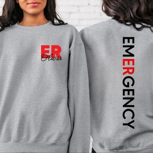 May include: Gray sweatshirt with the text "ER Olivia" in red and black on the front and "EMERGENCY" in black on the back.
