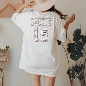 May include: White baseball-themed t-shirt with the name "MIKE" and the number "15" printed on the back. The lettering and numbers are designed to resemble baseballs, featuring red stitching details. The shirt is a casual, oversized fit.