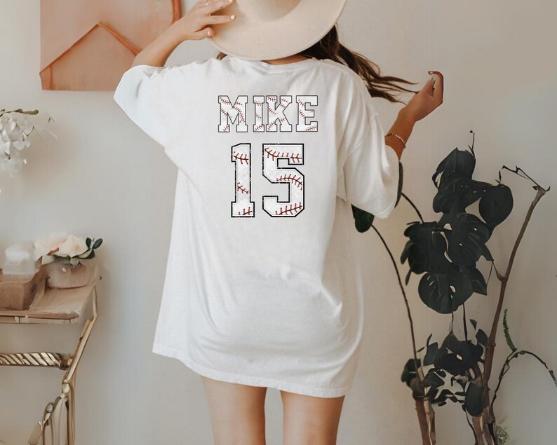 May include: White baseball-themed t-shirt with the name "MIKE" and the number "15" printed on the back. The lettering and numbers are designed to resemble baseballs, featuring red stitching details. The shirt is a casual, oversized fit.