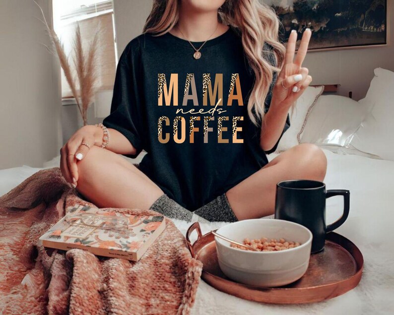 Op de afbeelding: Een zwart T-shirt met de tekst "MAMA needs COFFEE" in gouden letters met een luipaardprint. Het shirt wordt gedragen door een persoon die op een bed zit met een deken en een kom ontbijtgranen.