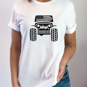 May include: White t-shirt with a black silhouette of a Jeep with an American flag on the hood and trees in the grill.