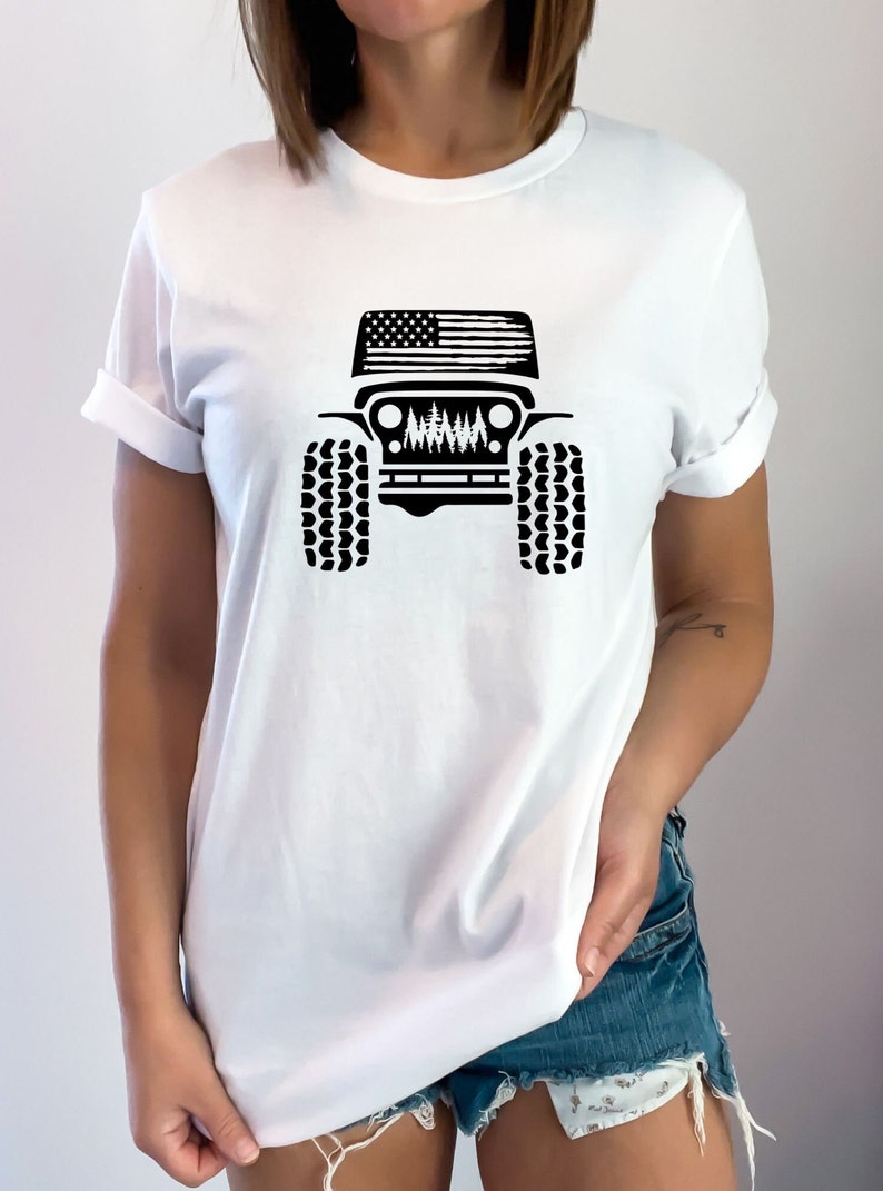 May include: White t-shirt with a black silhouette of a Jeep with an American flag on the hood and trees in the grill.