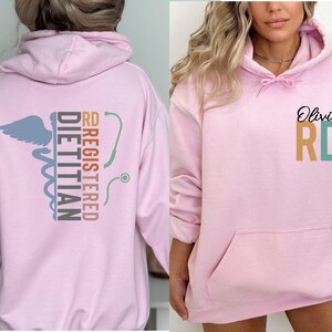 May include: A light pink hoodie with a front pocket and a hood. The back of the hoodie features the words "REGISTERED DIETITIAN" and "RD" with a caduceus symbol and a stethoscope. The front of the hoodie has the name "Olivia" and the letters "RD".