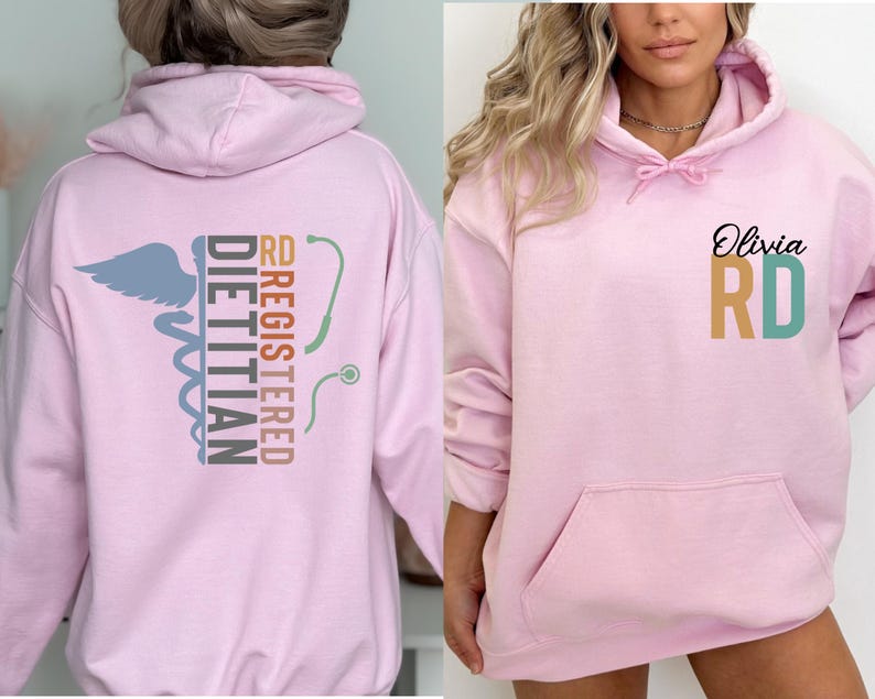 May include: A light pink hoodie with a front pocket and a hood. The back of the hoodie features the words "REGISTERED DIETITIAN" and "RD" with a caduceus symbol and a stethoscope. The front of the hoodie has the name "Olivia" and the letters "RD".