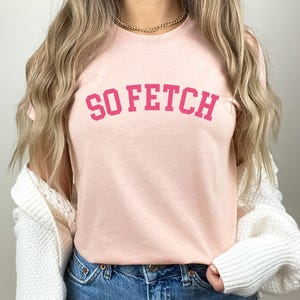 Op de afbeelding: Een roze T-shirt met de tekst "SO FETCH" in roze letters op de voorkant.
