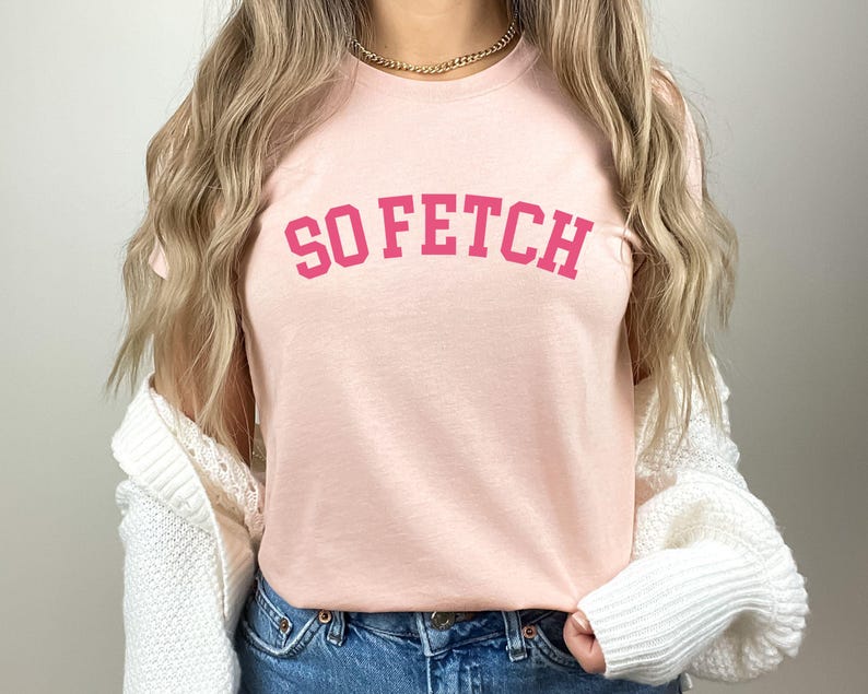 Op de afbeelding: Een roze T-shirt met de tekst "SO FETCH" in roze letters op de voorkant.